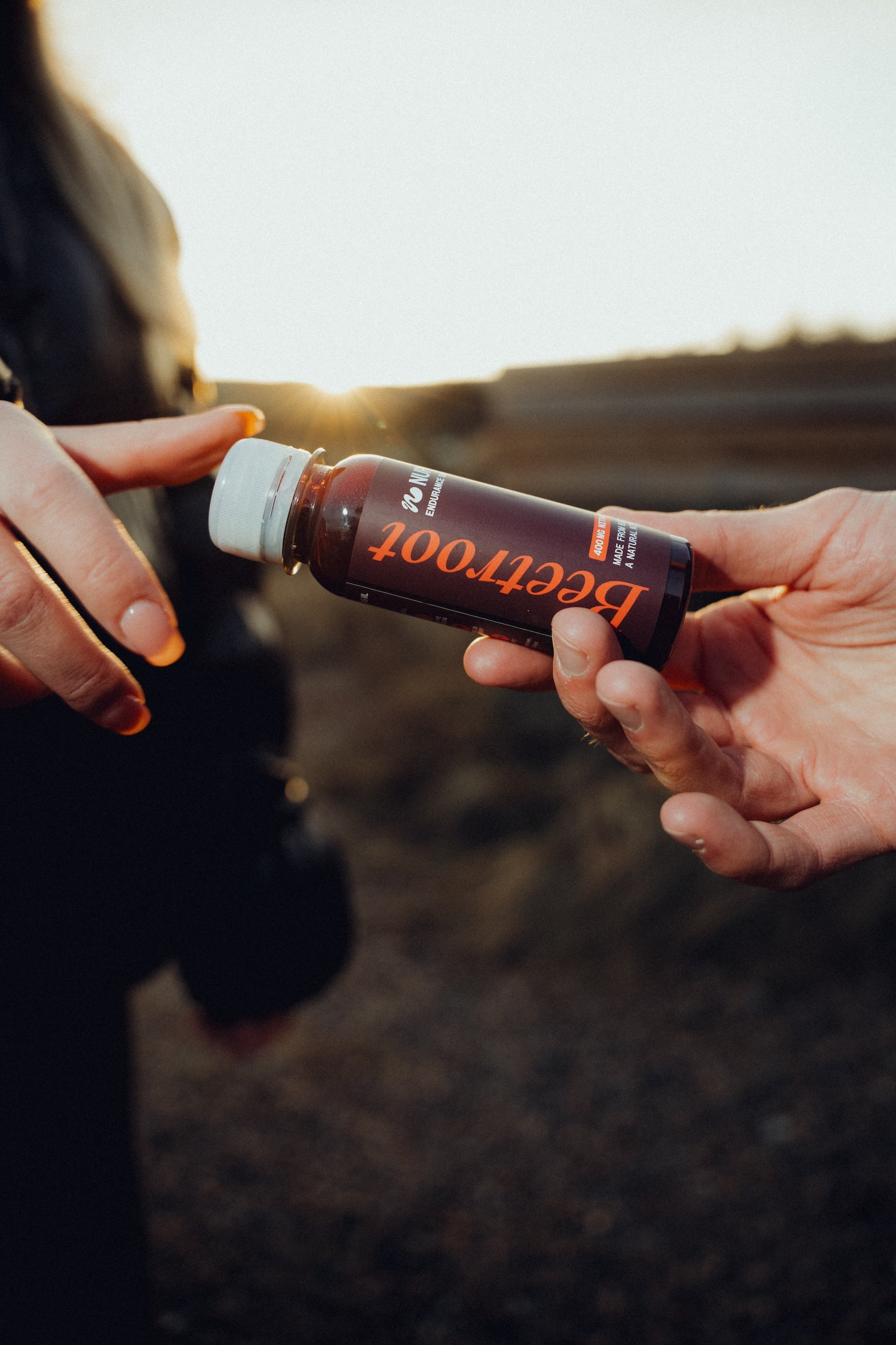 Hand passing a beetroot shot bottle outdoors at sunset, natural endurance supplement